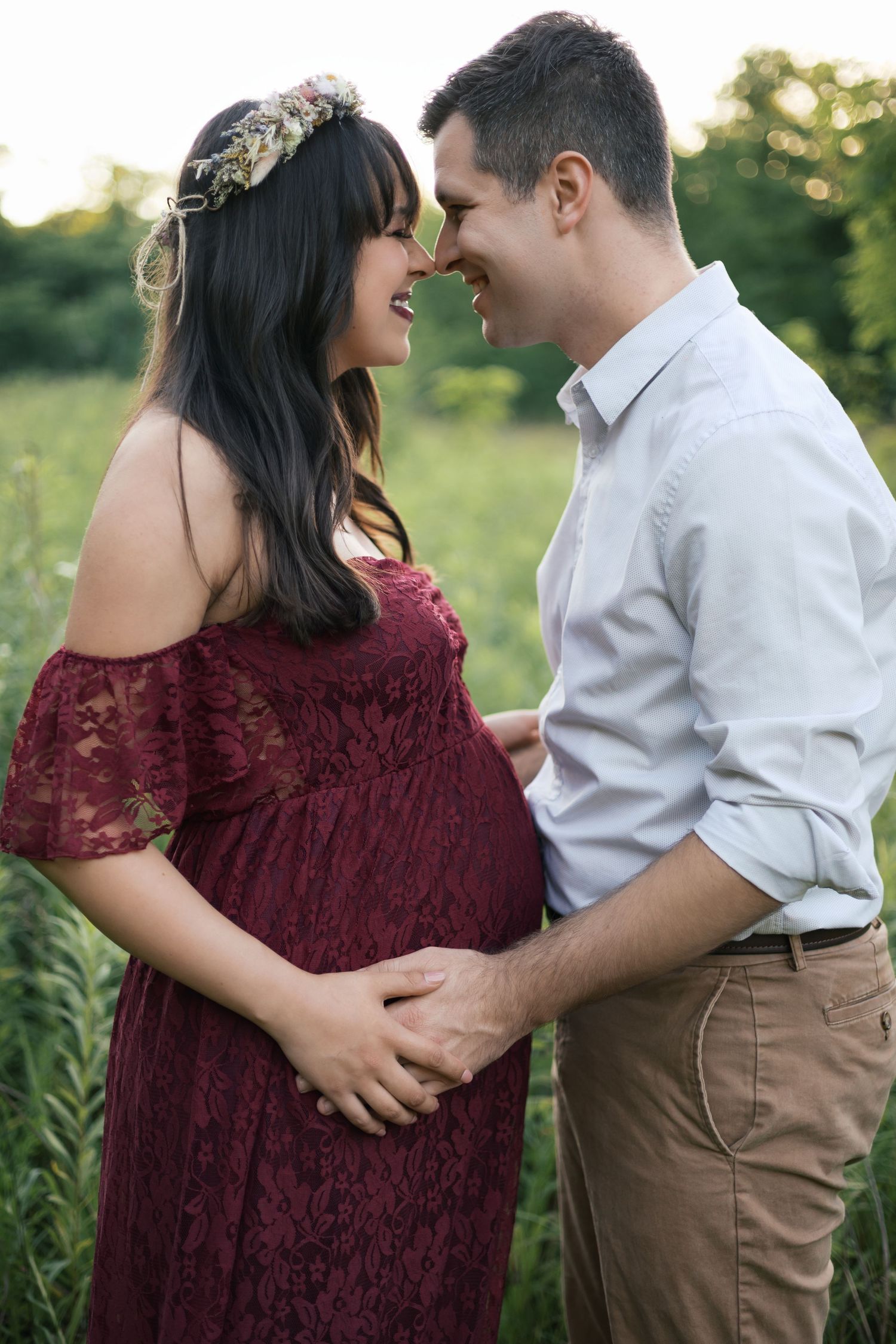 Pregnant couple in a green field, woman in red lace dress with floral crown, man in white shirt, smiling at each other.