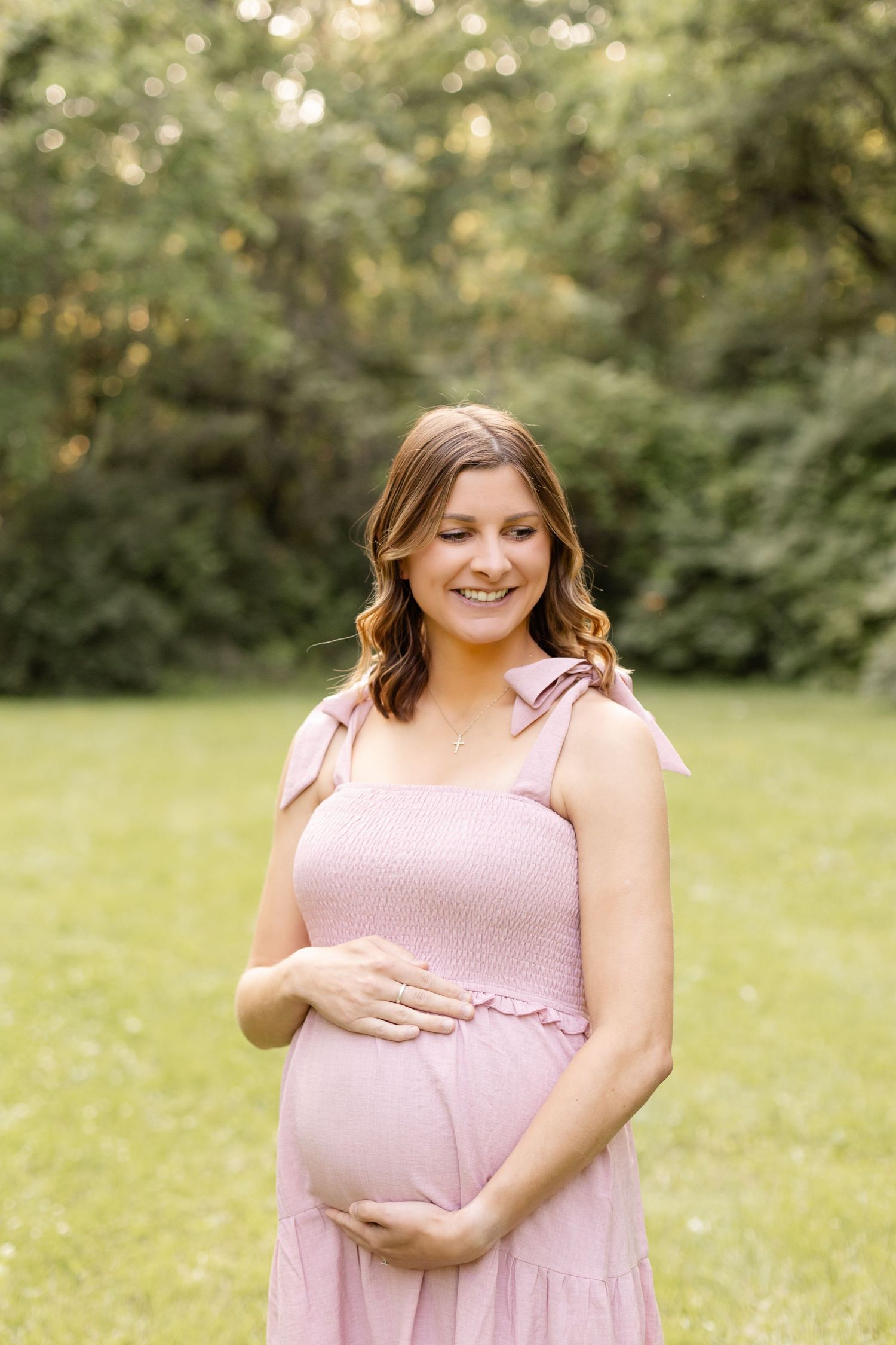 Expectant mother in pink dress poses in sunny garden setting, gently cradling baby bump.