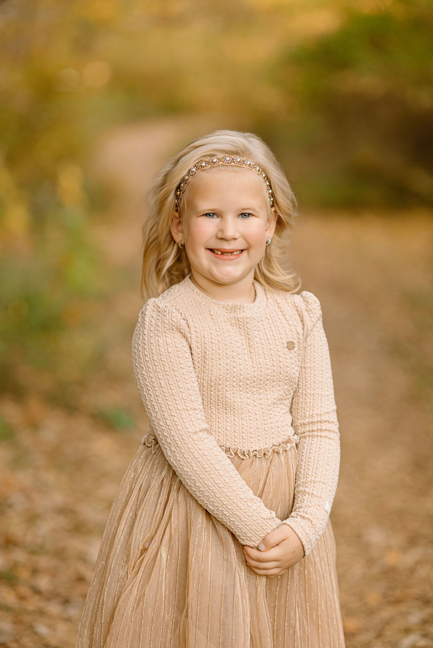 A smiling blonde girl in a beige knit dress and headband poses on a sunlit autumn dirt path.