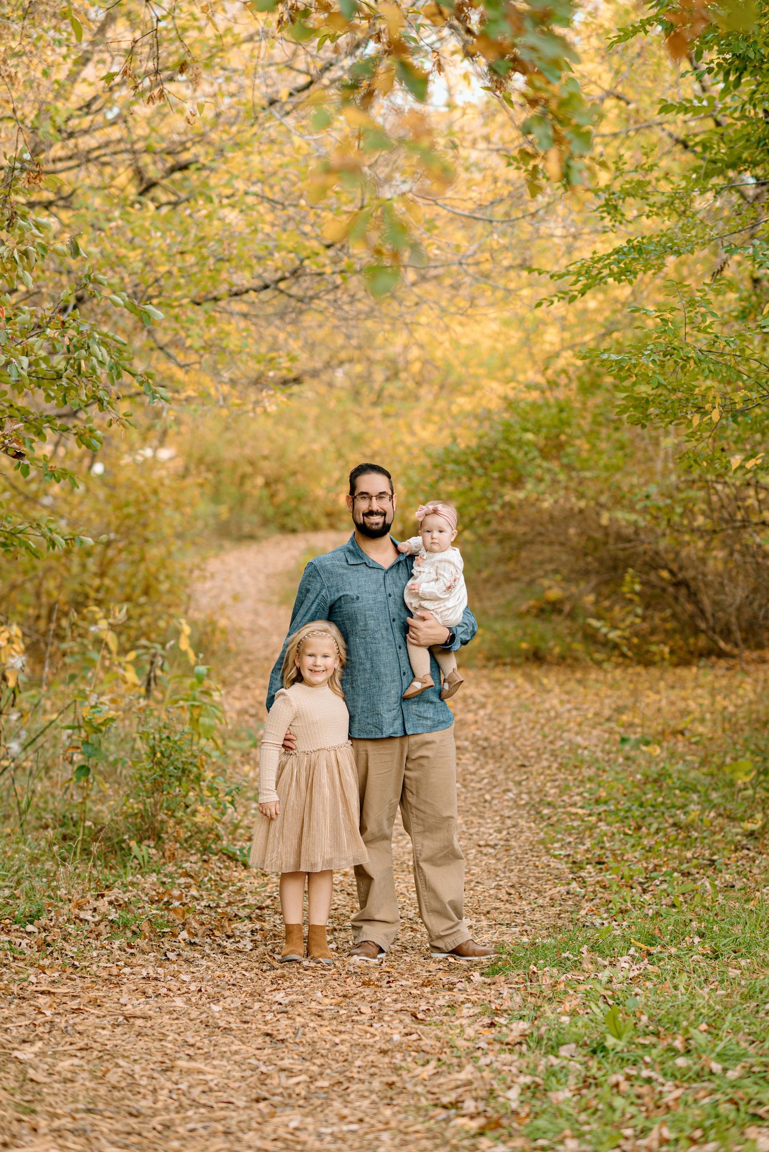 A bearded father stands on a fall nature path holding a baby while his young daughter stands beside him.