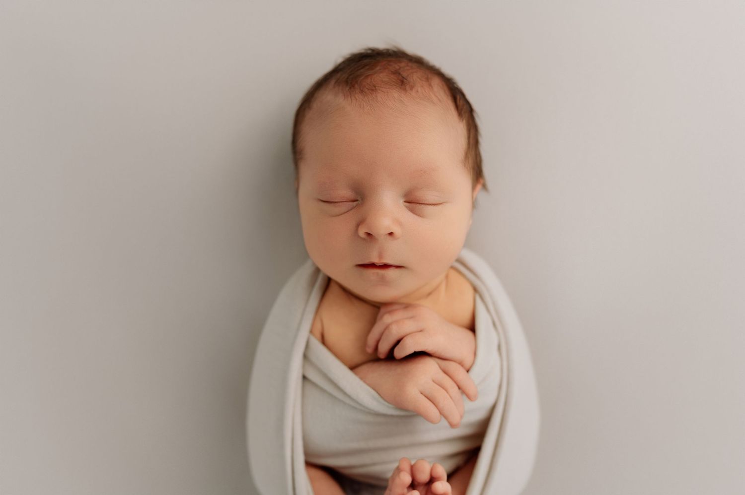 Newborn baby wrapped in soft gray swaddle with hands folded, sleeping peacefully on gray backdrop.