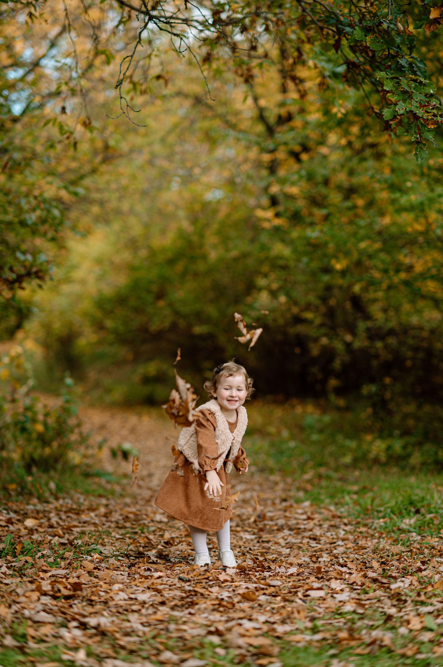 Joyful toddler girl in a brown dress laughs while tossing autumn leaves on a forest trail.