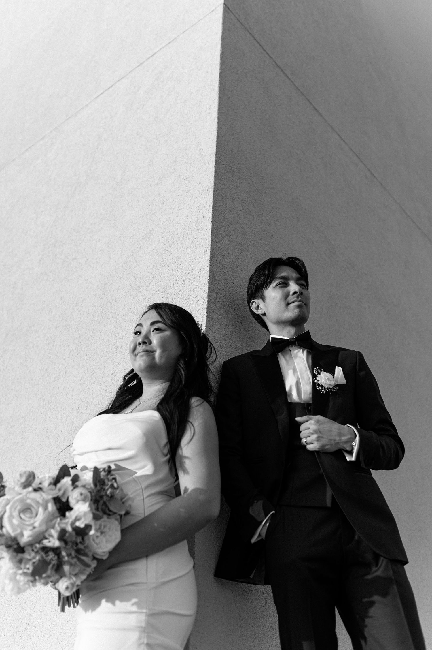 Black and white portrait of elegant bride and groom leaning against a textured wall.