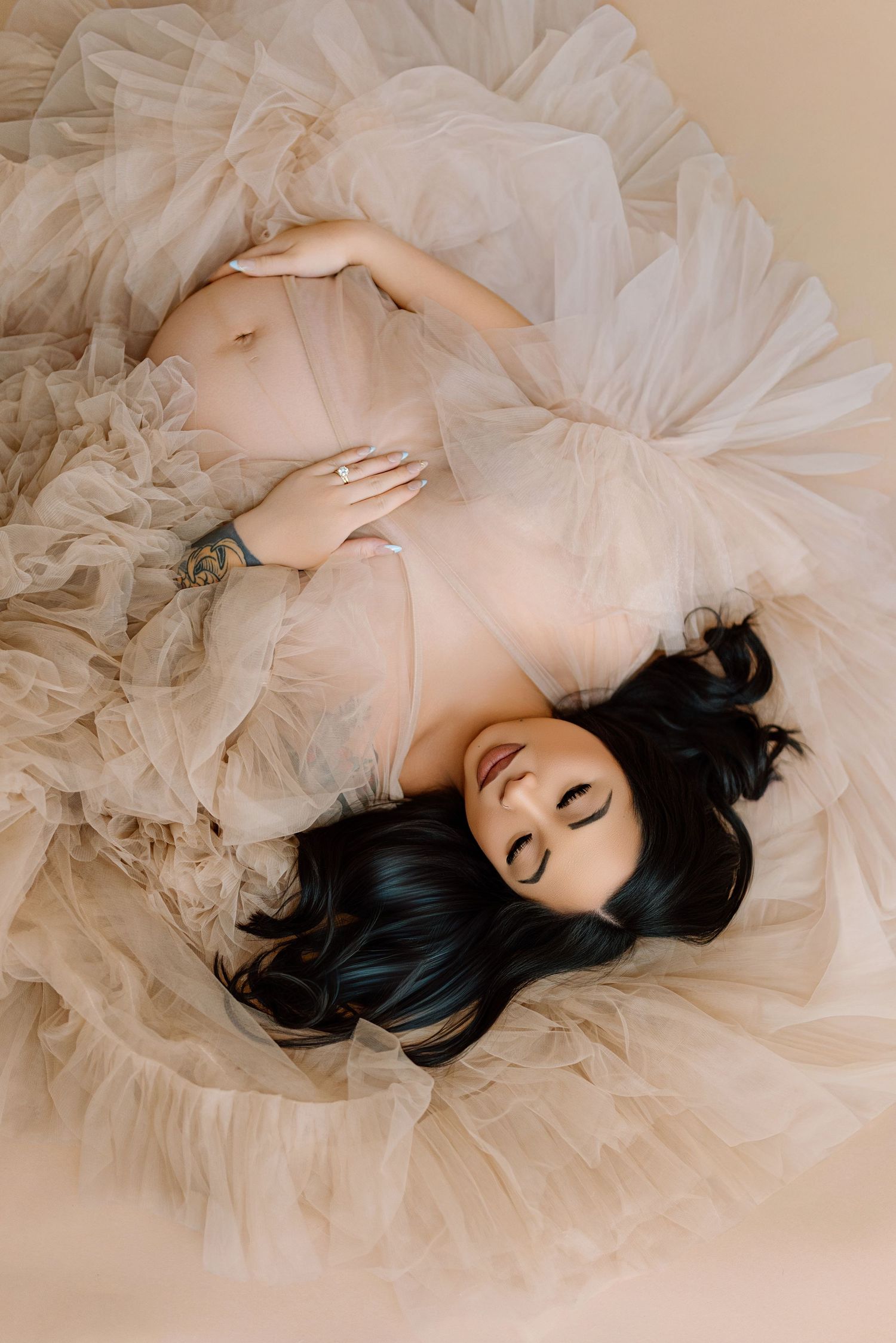 Expecting mother with black hair reclines gracefully in sheer beige tulle dress for maternity shoot.
