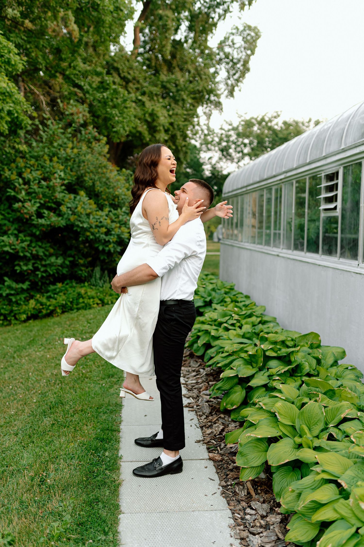 Groom lifts laughing bride in white dress outdoors beside greenhouse and lush green plants.