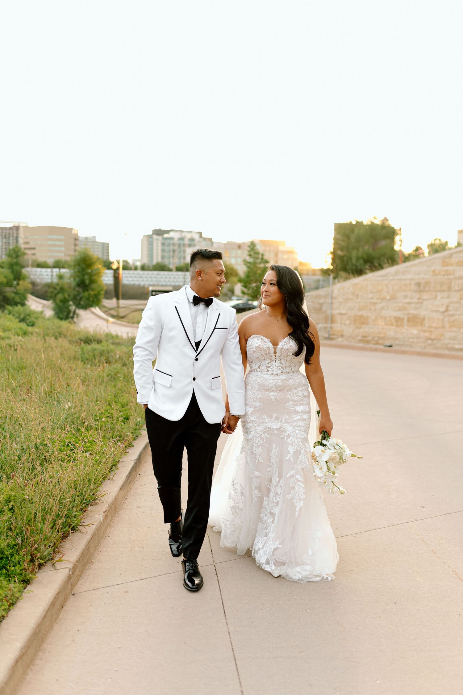 Couple walks at sunset, groom in white tuxedo jacket, bride in lace mermaid gown with bouquet.