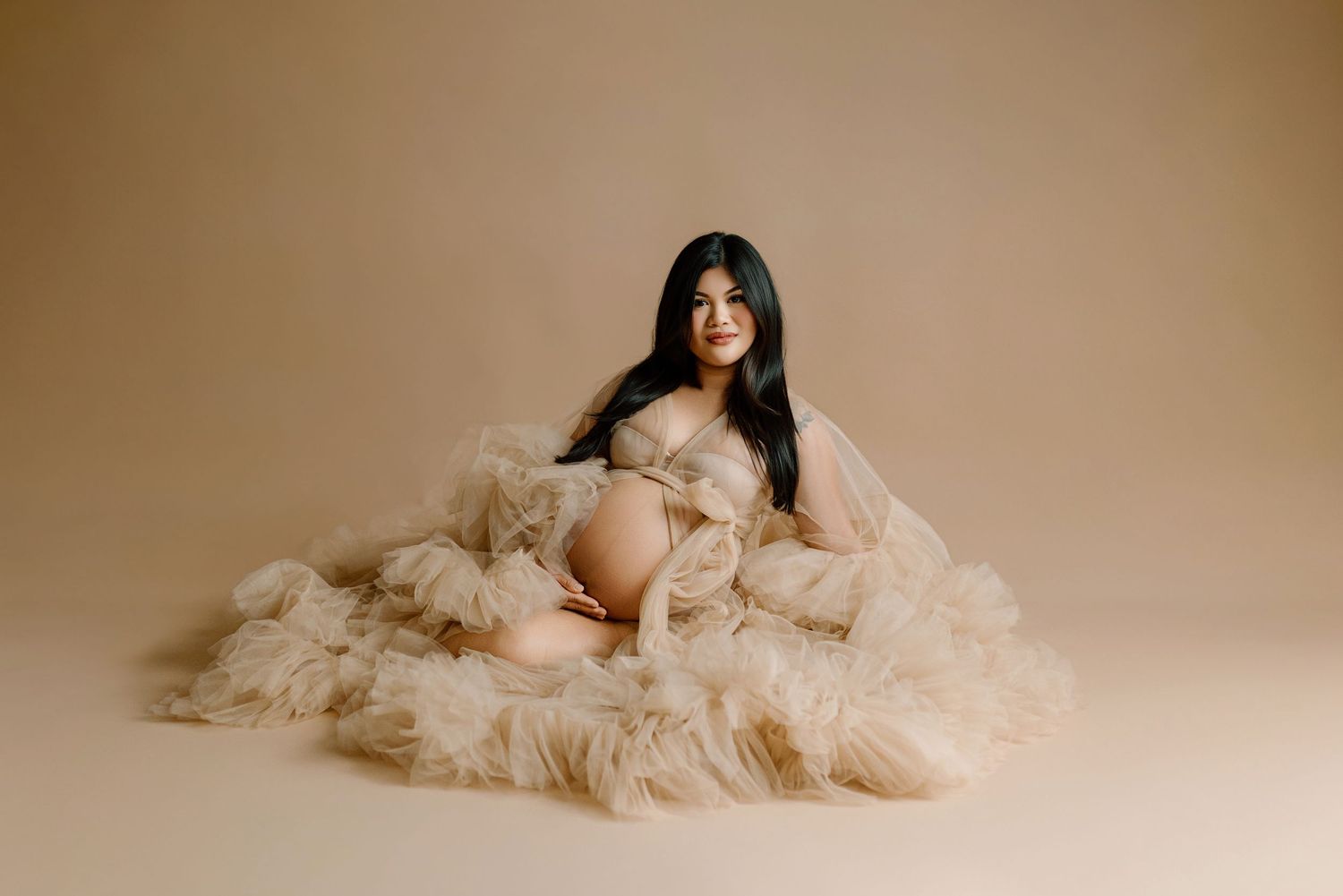 Pregnant woman in flowing beige tulle robe poses gracefully on studio floor against neutral background.
