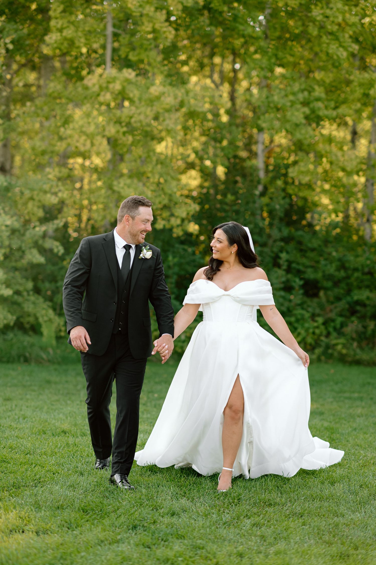 Bride and groom walk hand-in-hand across green lawn with golden autumn trees.