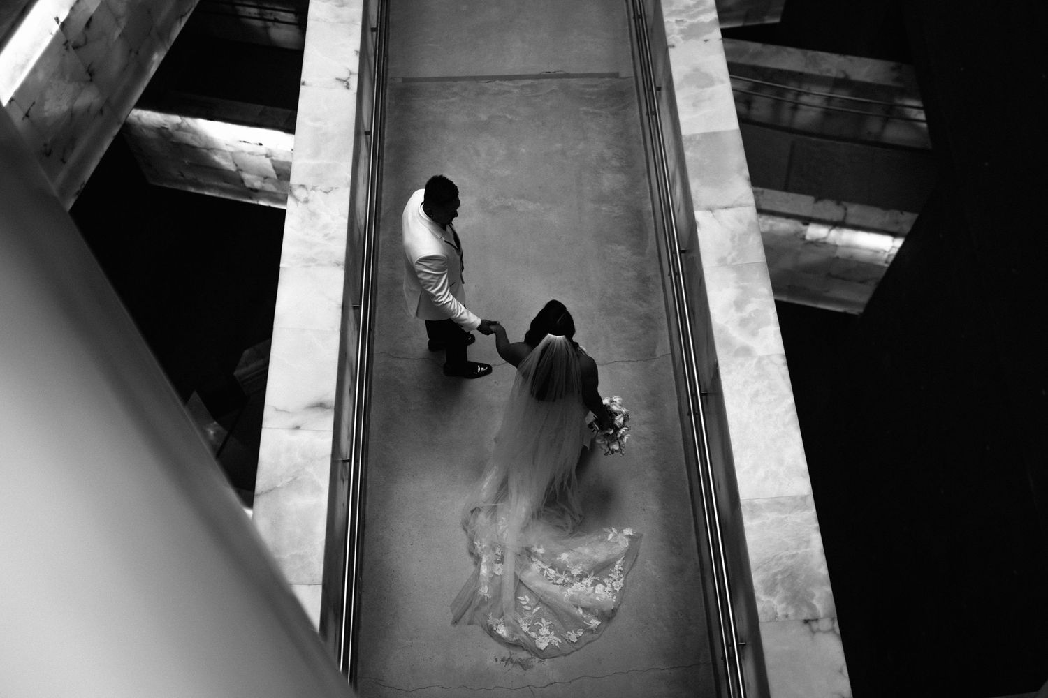 Black and white aerial view of bride and groom holding hands walking through a modern architectural corridor.