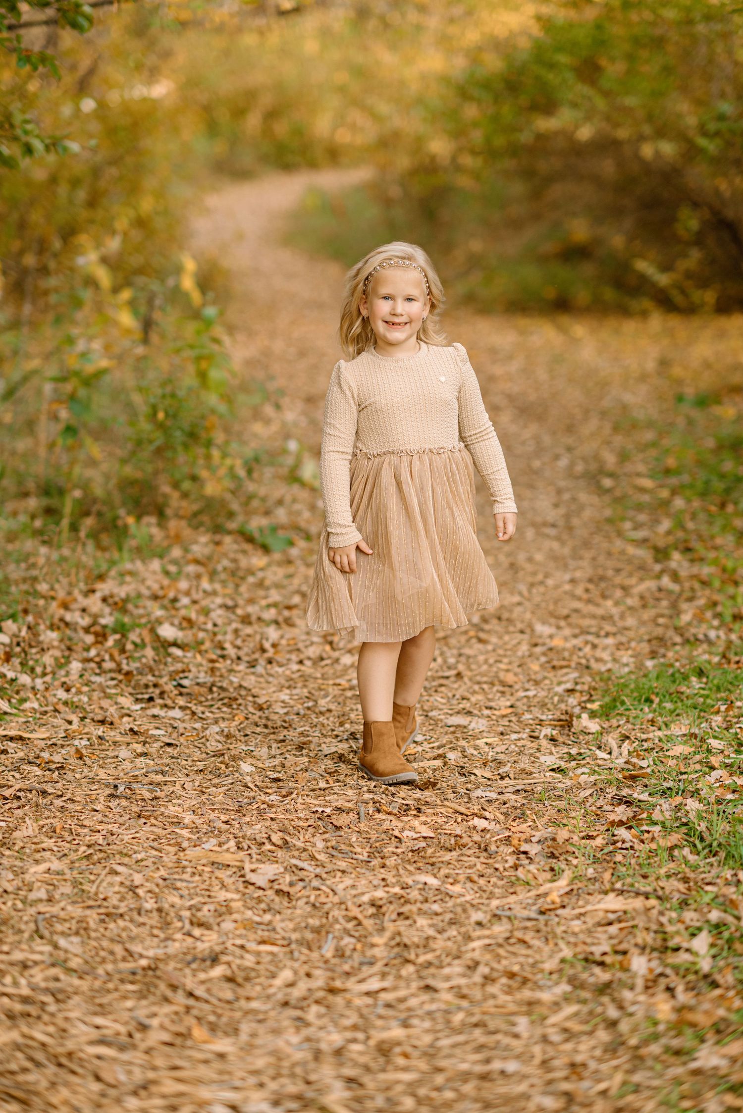 Young girl in beige dress and boots walking on autumn leaf-covered trail.