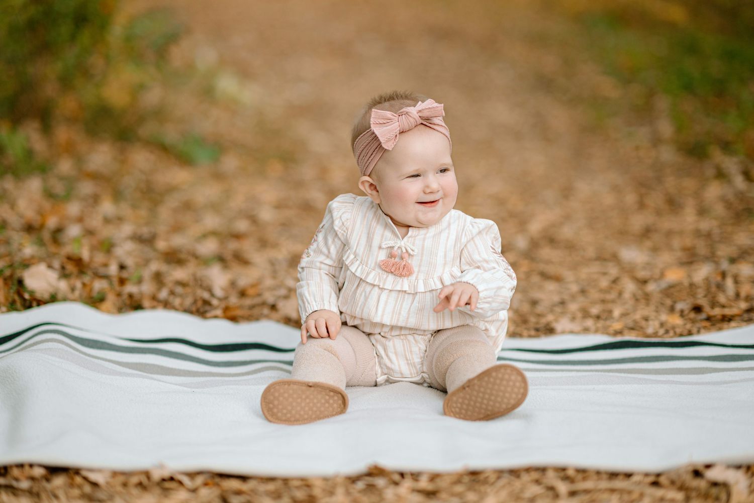 Smiling baby girl in cream romper and pink bow headband sitting on white blanket among autumn leaves outdoors.