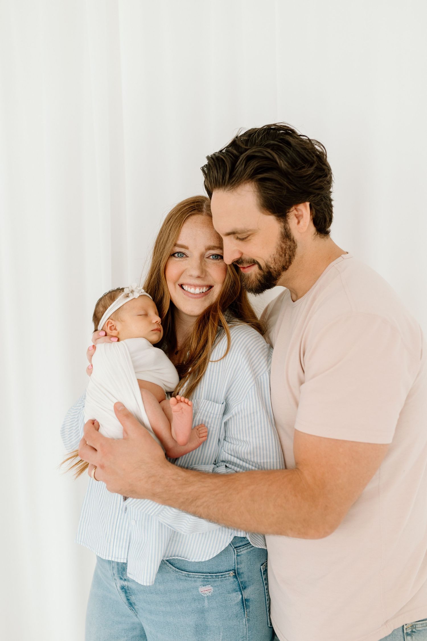 Happy new parents in casual clothes holding their swaddled newborn baby girl with headband.