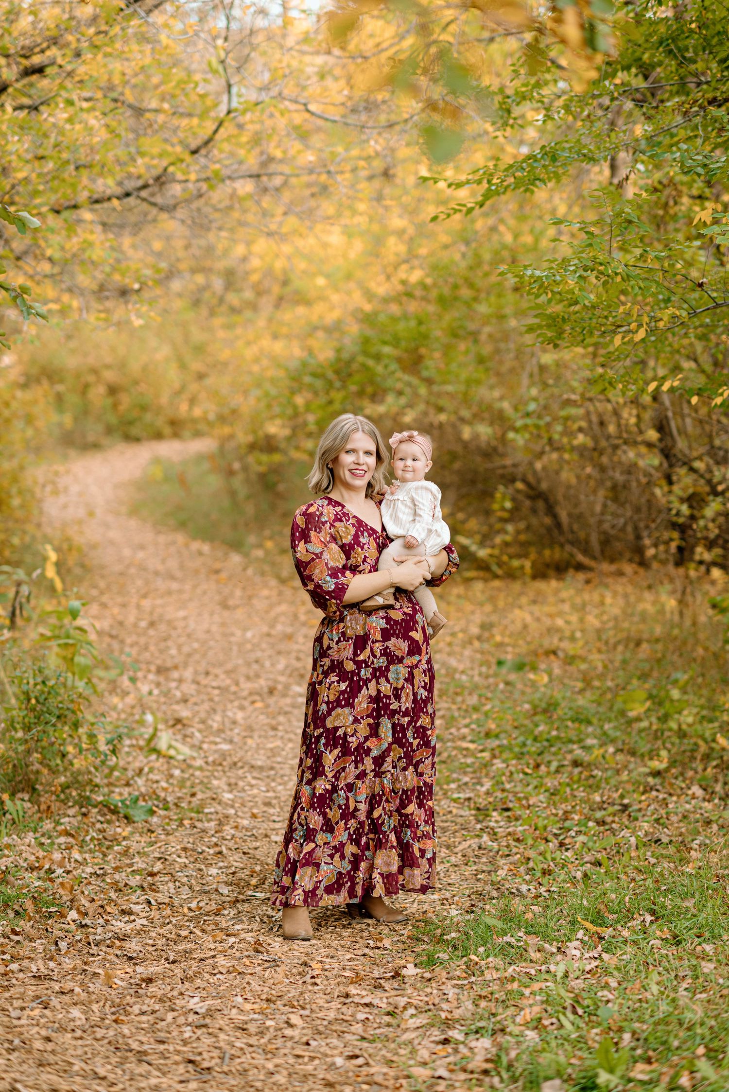A woman in a floral maxi dress holds a baby on a scenic autumn path lined with colorful trees.