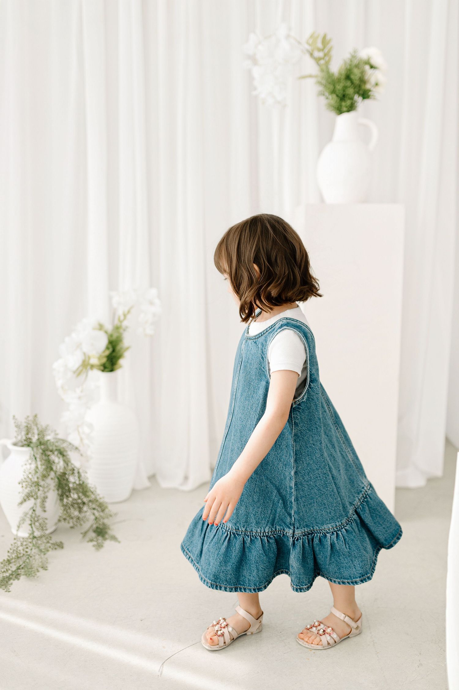 Young girl in denim dress walking in a bright white studio decorated with green plants and flowers.