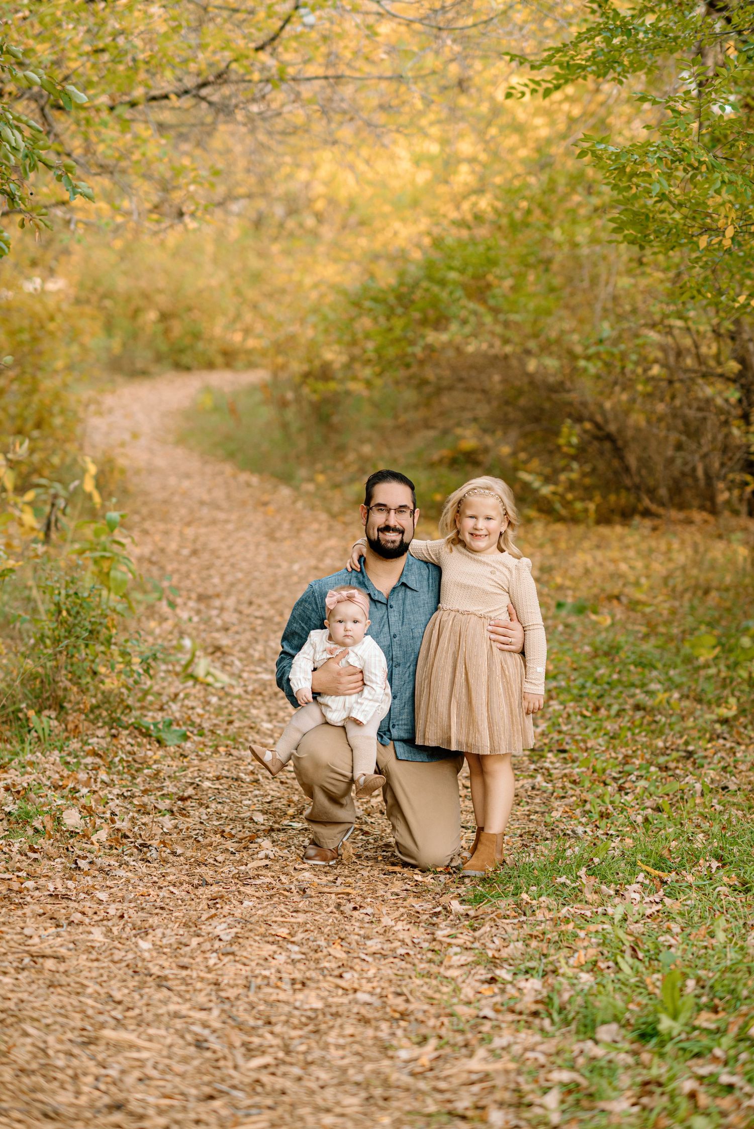 A bearded father kneels on a fall trail holding a baby while his young daughter stands beside him.