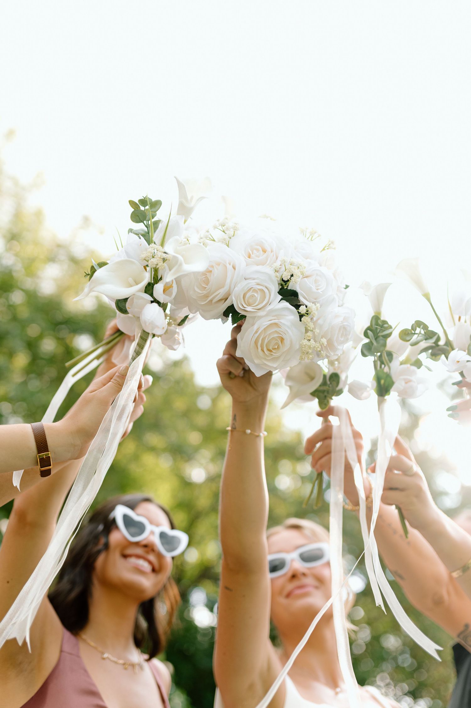 Bridesmaids joyfully raise white rose bouquets with ribbons toward bright sky.