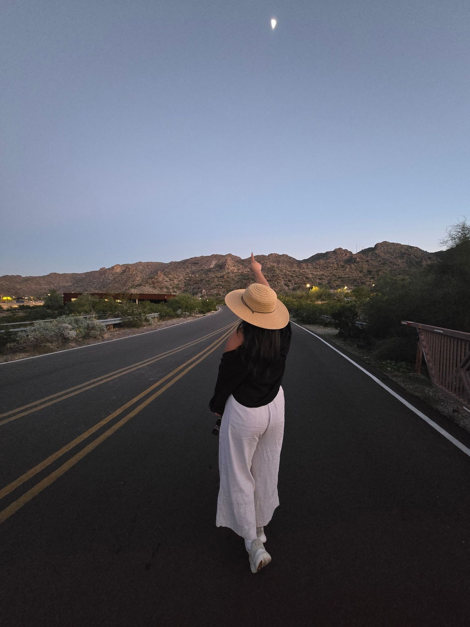 Woman in white pants and straw hat pointing at the moon on a desert road at dusk.