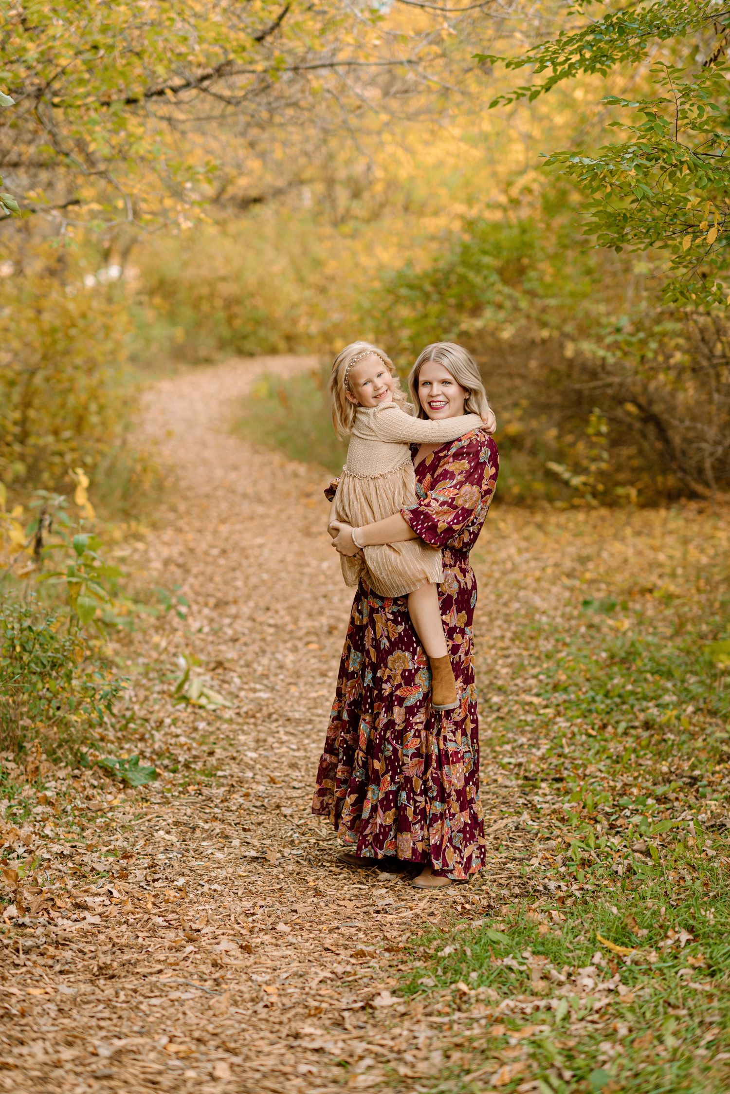 Woman in floral maxi dress holding young child on a leaf-covered autumn path surrounded by golden foliage.