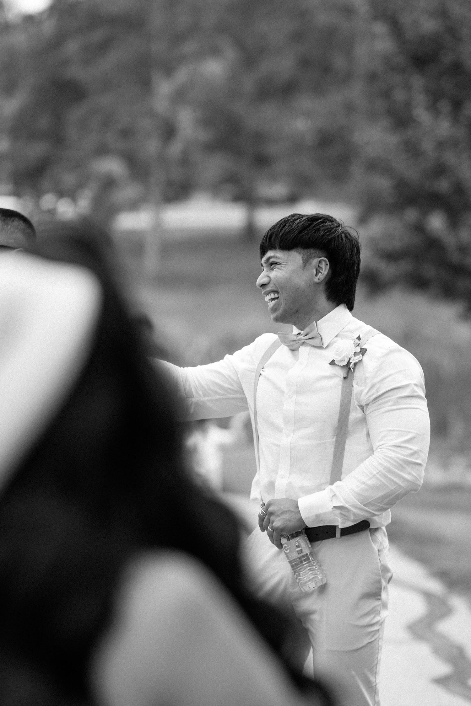 Smiling Asian groom in white shirt with suspenders and bow tie at outdoor wedding celebration.