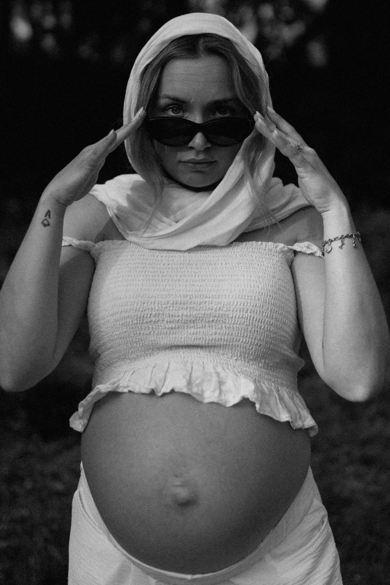 Pregnant woman in white crop top and hood poses confidently wearing sunglasses in black and white photo.