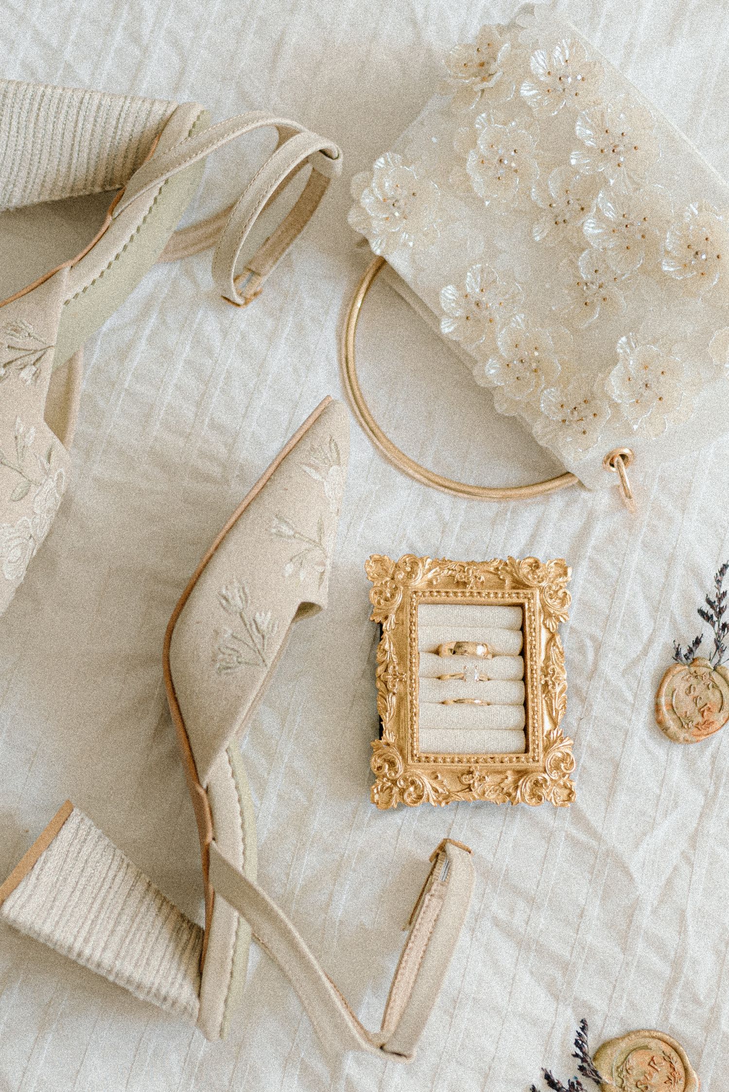 Elegant bridal flat lay with cream embroidered heels, gold ring frame, lace clutch, and delicate jewelry on white linen.