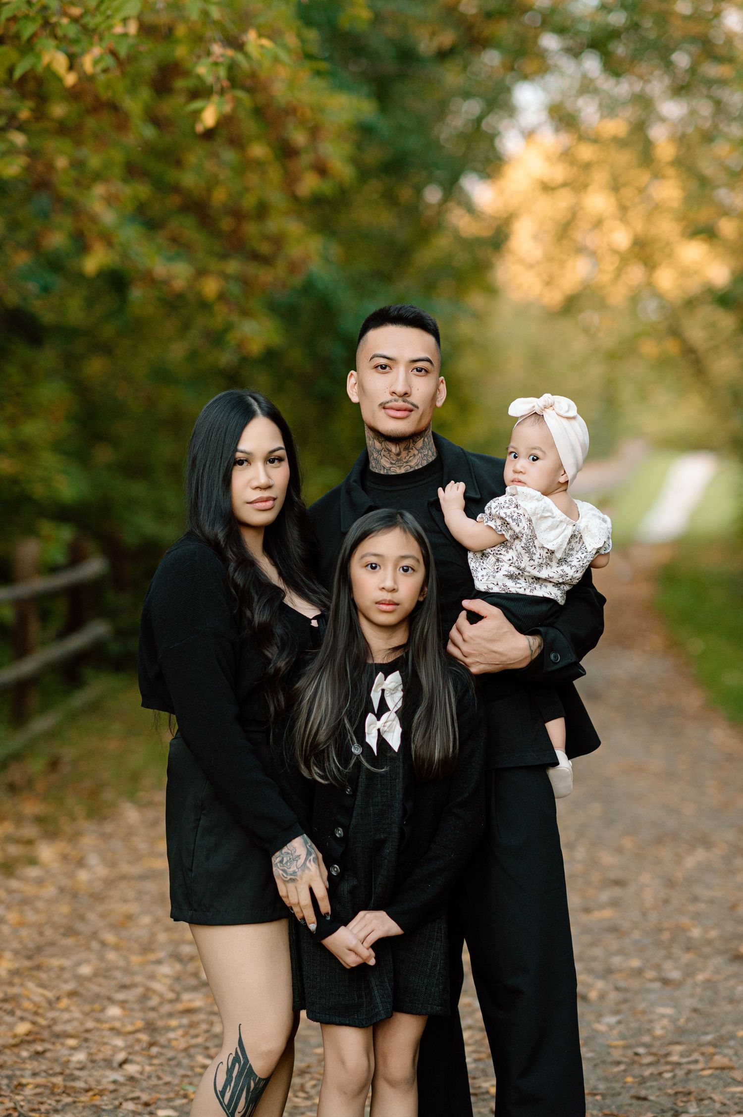 Family of four poses in matching black outfits on a tree-lined autumn path.