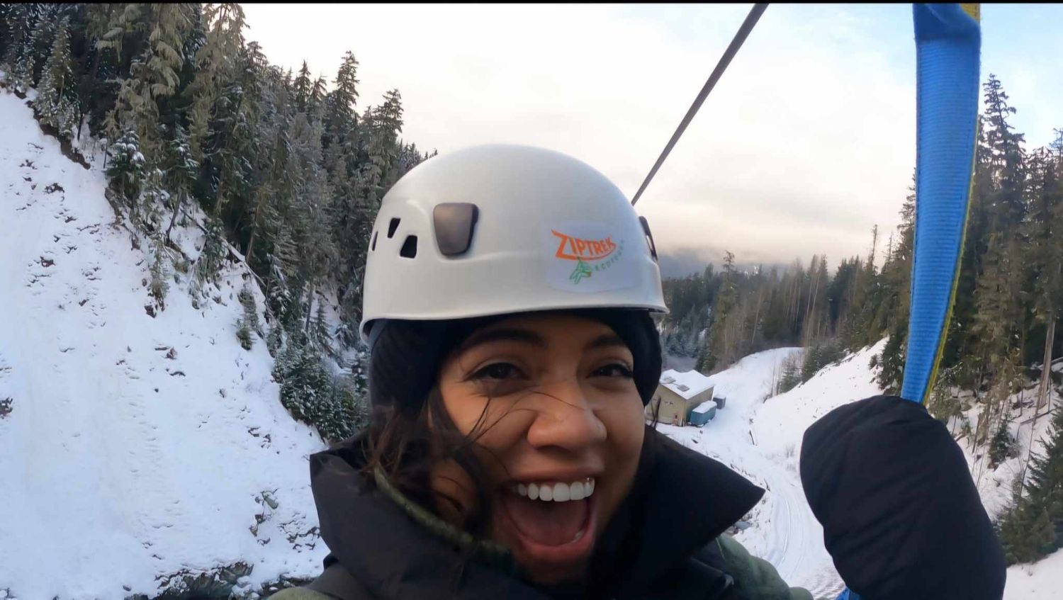 Excited woman zip lining through snowy forest wearing safety helmet outdoors.