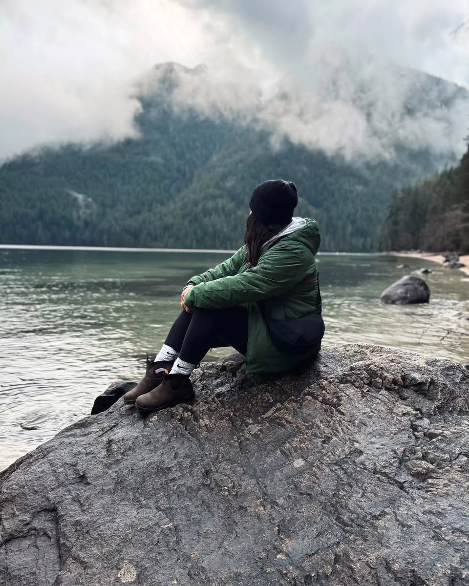 Woman in green jacket sitting on rocky shore with misty mountain lake backdrop.