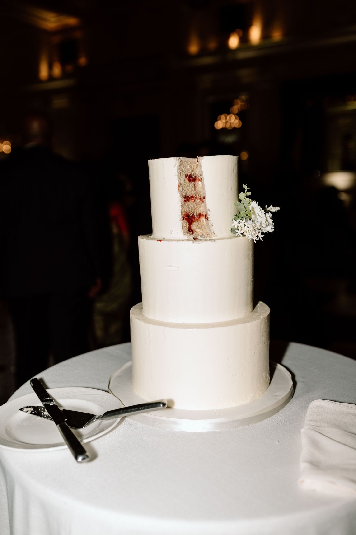 Elegant three-tier white wedding cake with a slice removed revealing red berry filling, topped with white flowers.