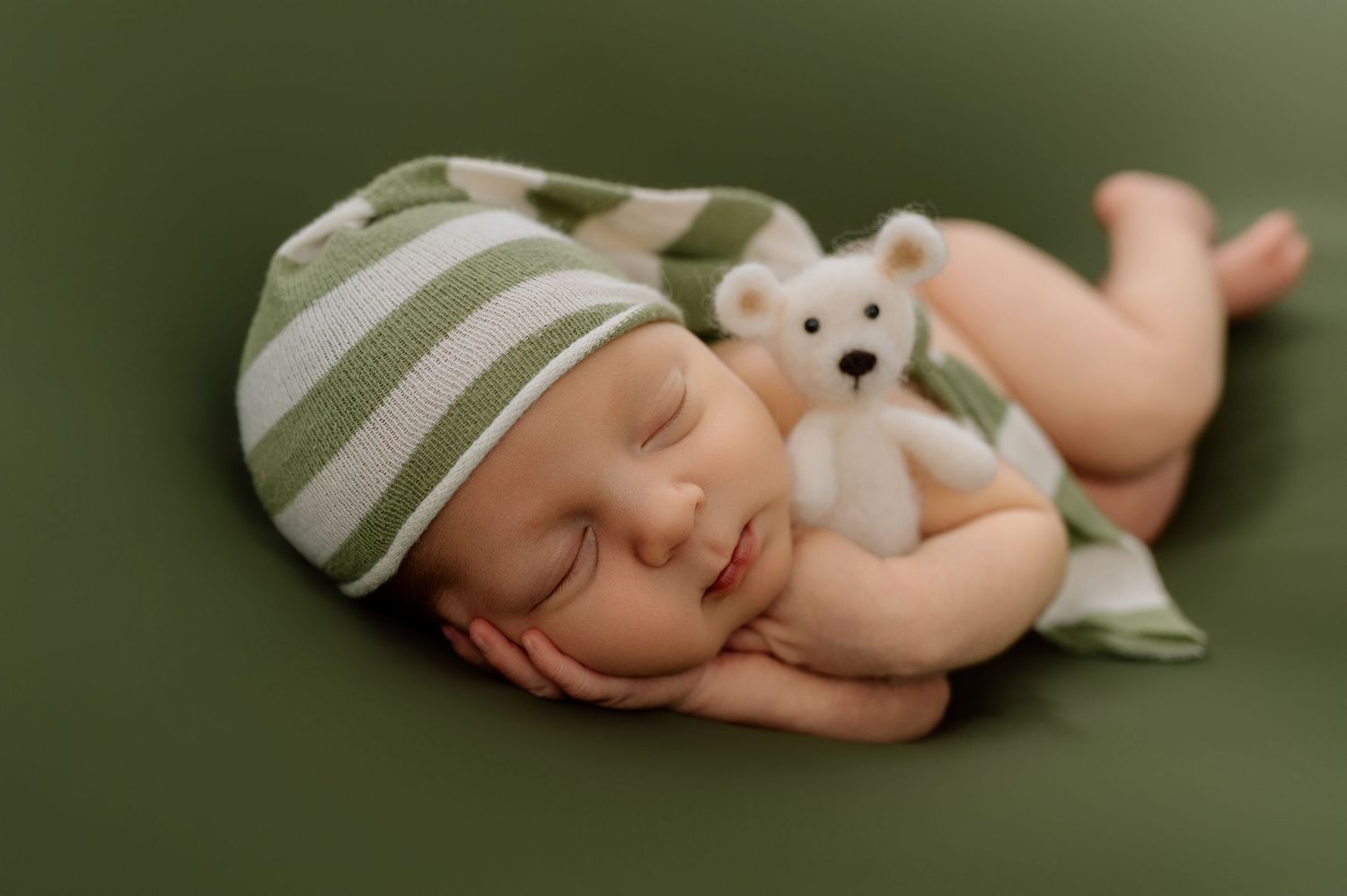 Sleeping newborn baby wearing striped green hat cuddling white teddy bear on green backdrop.