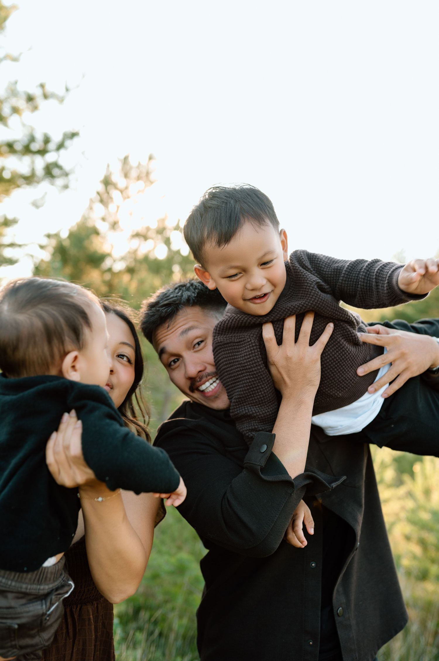 Happy family with two young boys playing outdoors in a sunlit forest setting.