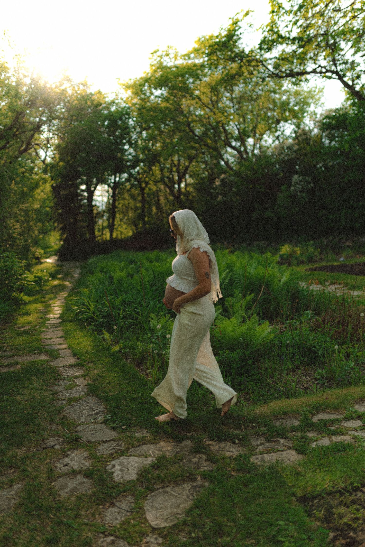 Pregnant woman in white outfit and headscarf walks along stone garden path surrounded by lush greenery at sunset.