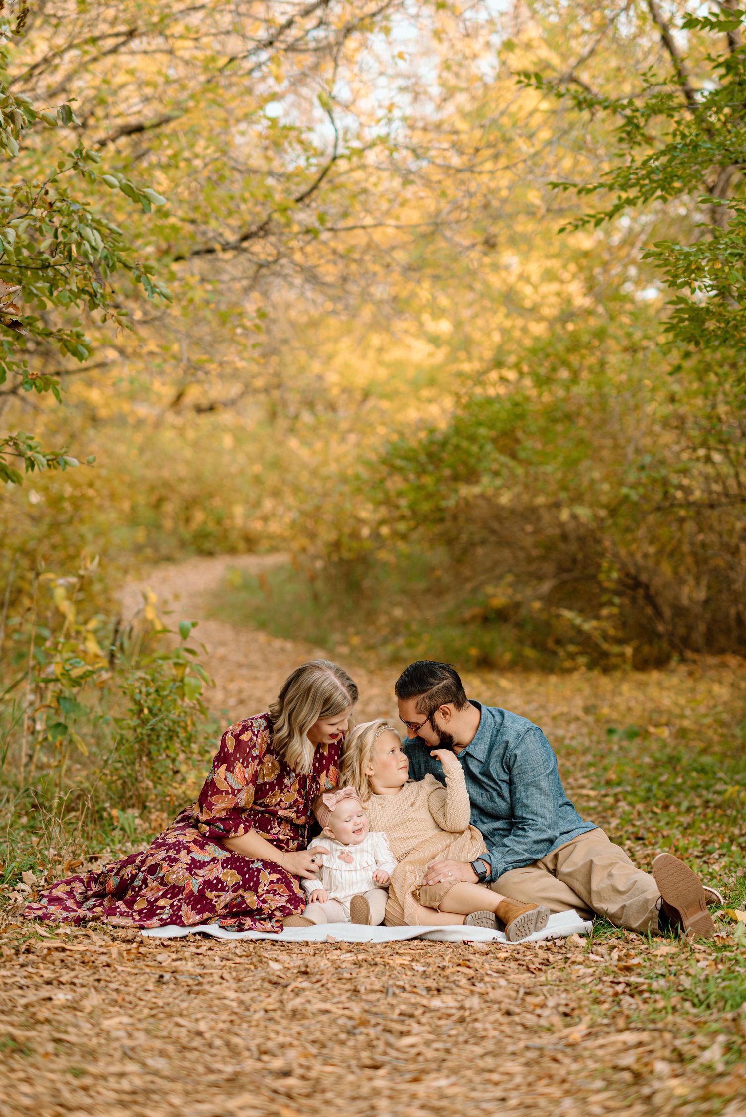 Family of three sitting on a blanket surrounded by golden autumn foliage on a scenic forest path.