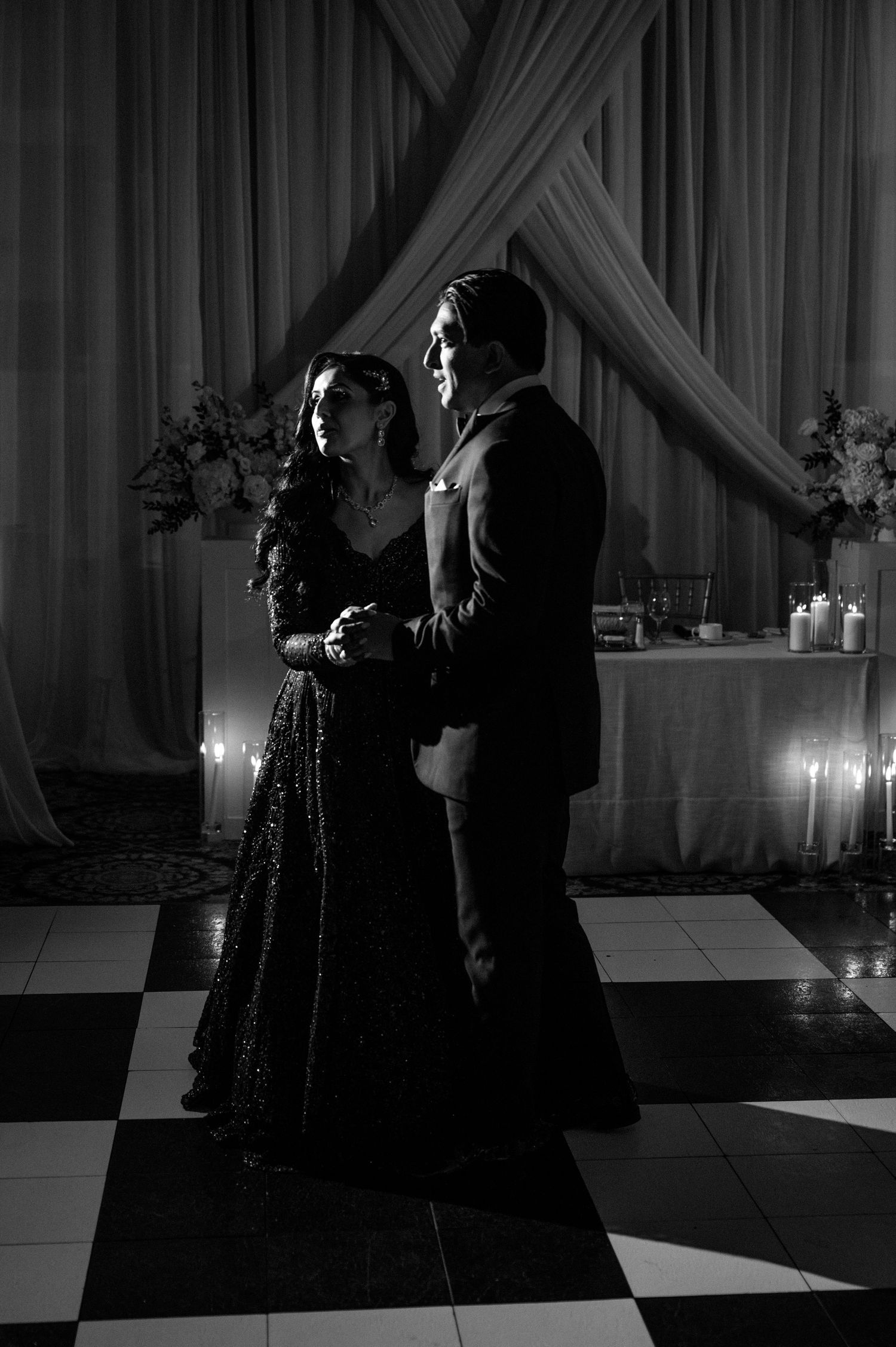 Romantic black and white photo of couple sharing first dance on checkered floor with candlelight.