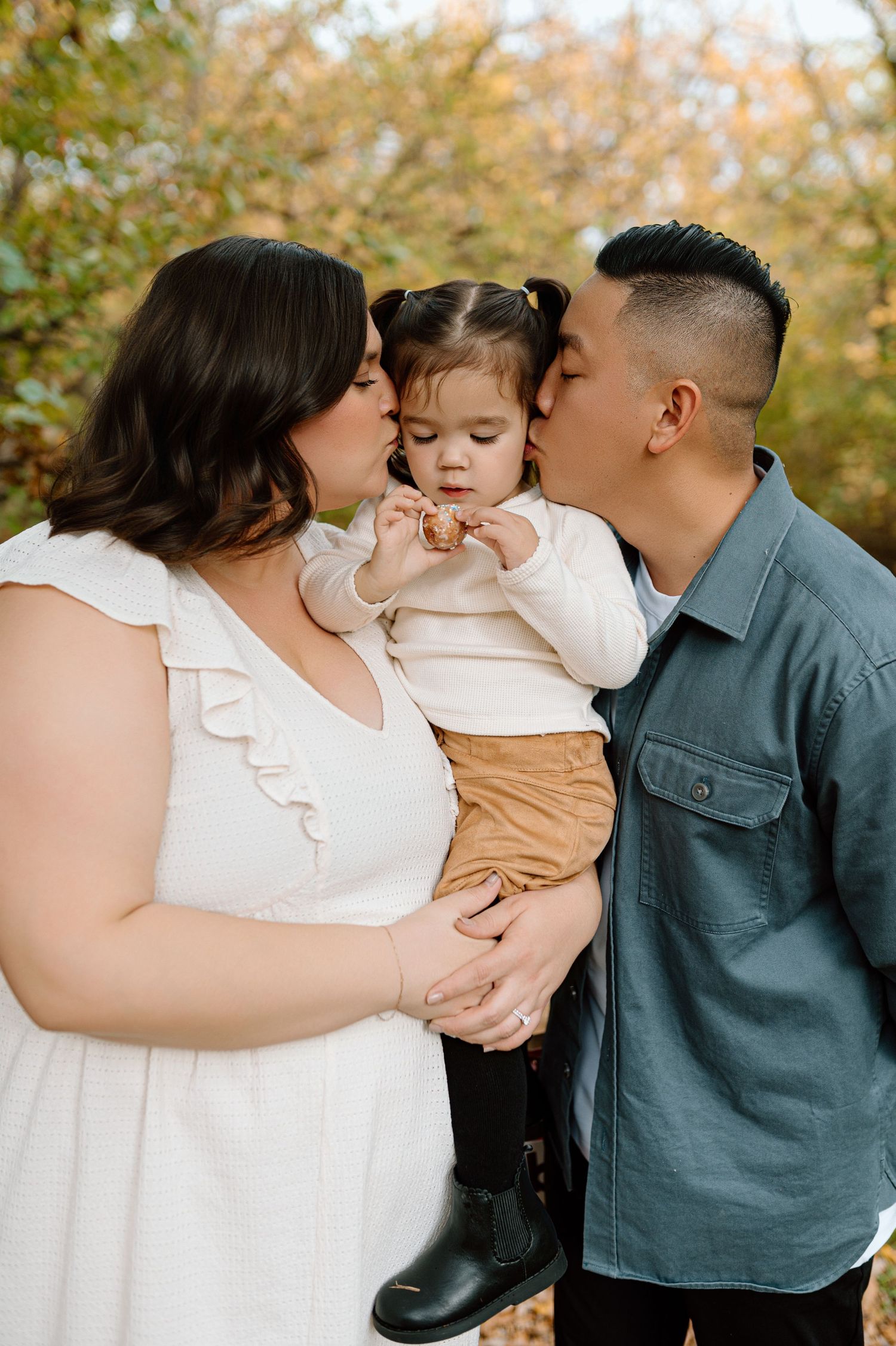Parents lovingly kiss their toddler daughter on the cheeks during an outdoor fall family portrait.