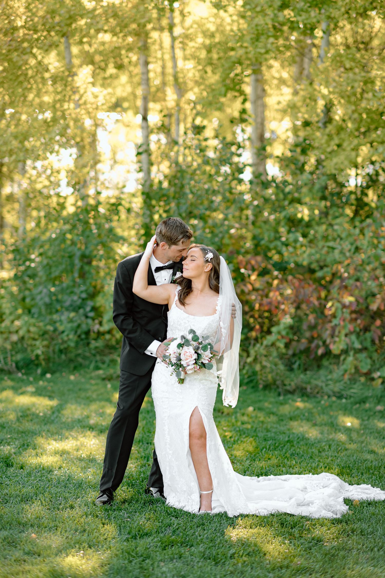 Groom tenderly kisses bride holding floral bouquet in front of sunlit aspen trees.
