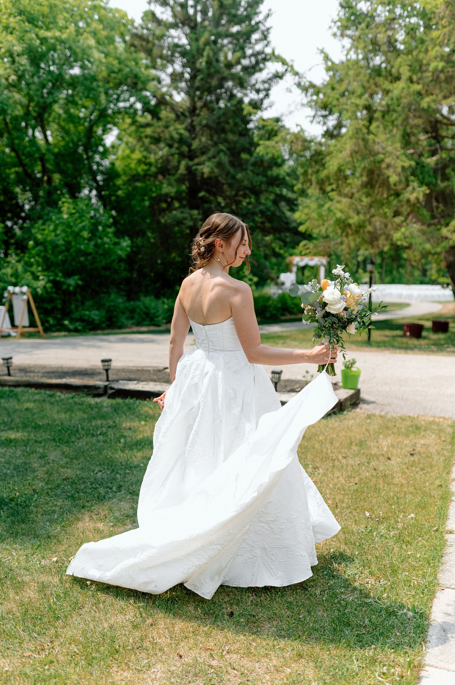 Bride in strapless white ballgown twirls holding bouquet of peach and white flowers outdoors.