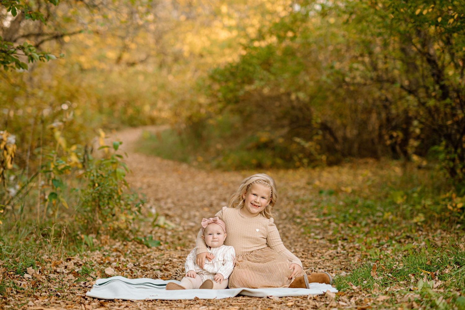 Two young sisters sit together on a blanket along a leaf-covered path surrounded by golden autumn foliage.