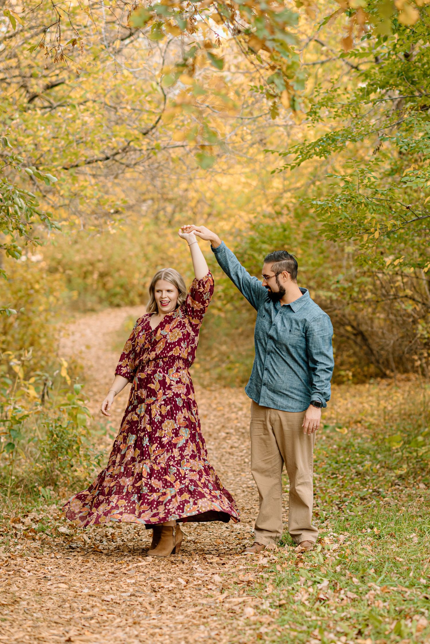 Couple dancing on a leaf-covered autumn path, woman in floral burgundy dress, man in blue shirt holding her hand up.