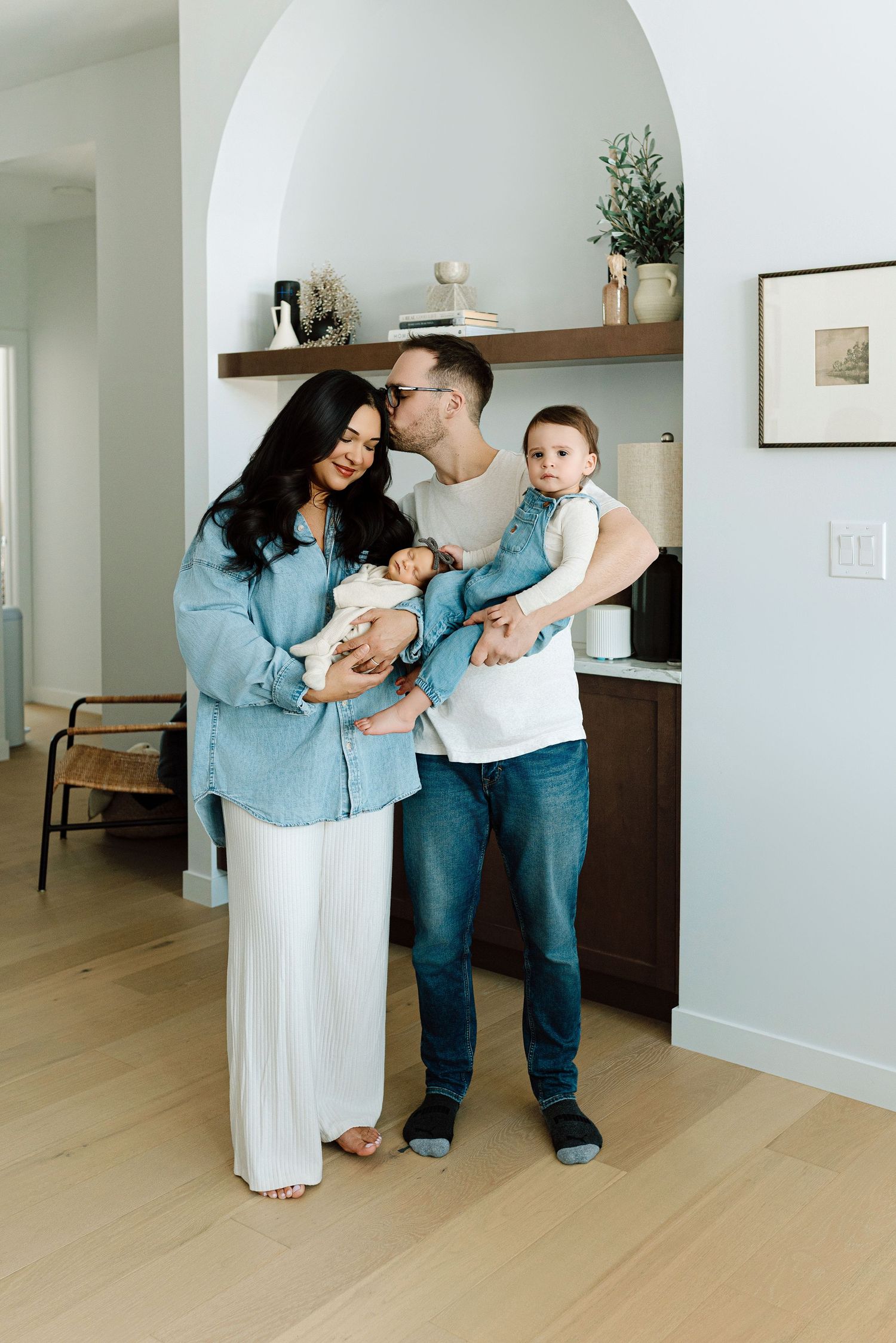 Young family with newborn and toddler standing under an arched alcove in a modern bright home.