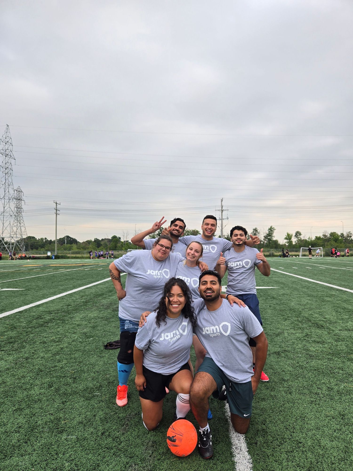 Group of seven friends posing on green turf field wearing matching gray team jerseys during outdoor sports event.