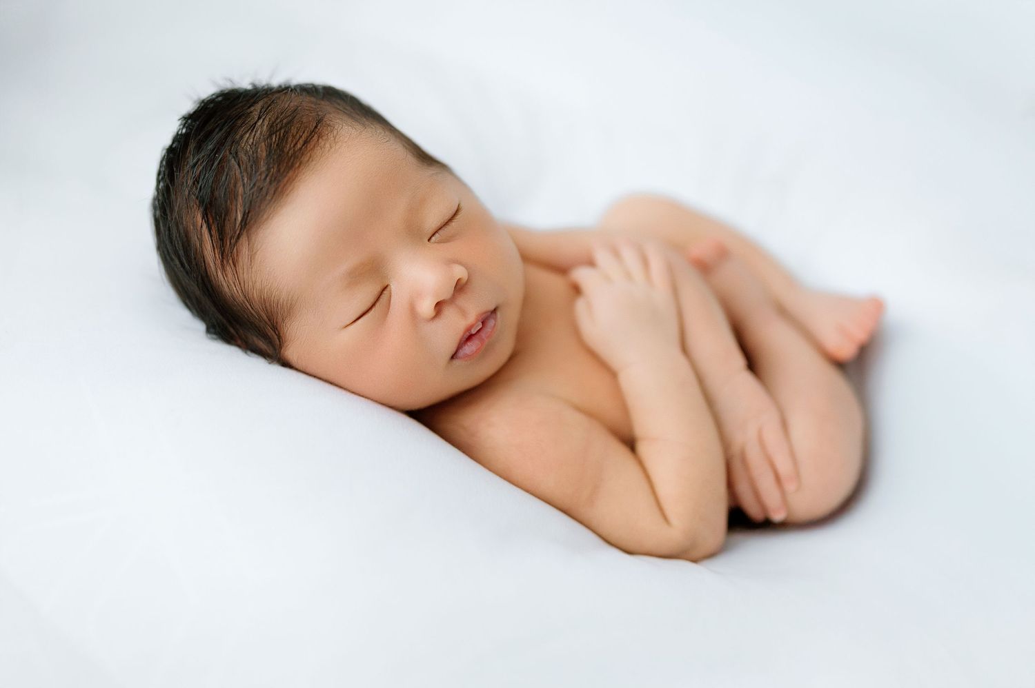Peaceful newborn baby sleeping on white blanket with hands tucked under chin in natural pose.