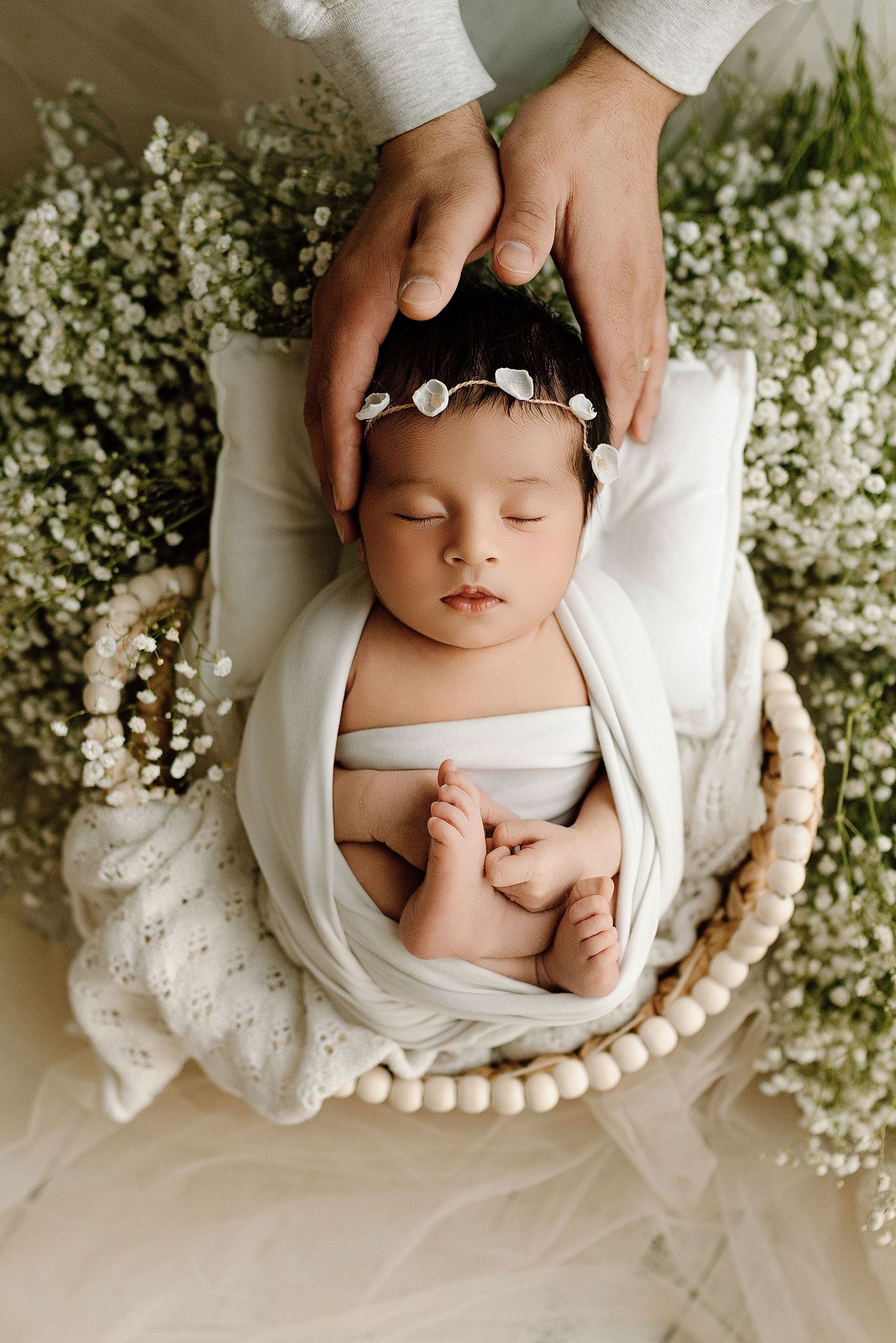 Sleeping newborn baby wrapped in white swaddle with floral headband in a basket surrounded by baby's breath flowers.