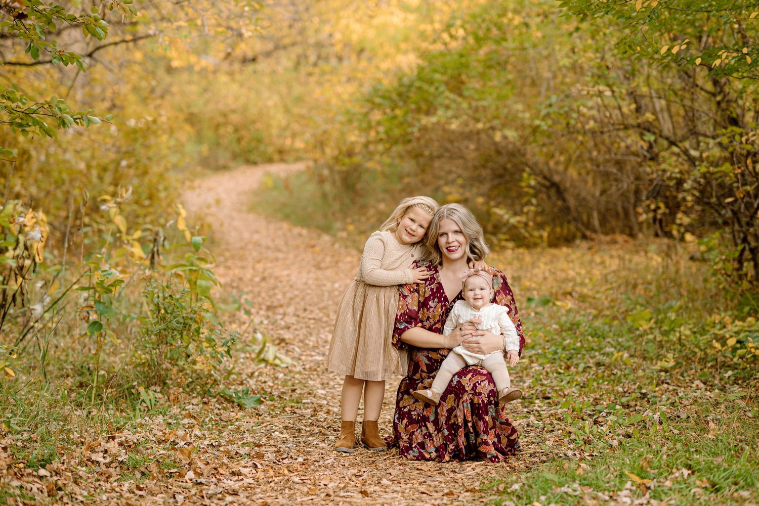 A mother kneels on a leaf-covered path holding a baby while a young girl hugs her in an autumn forest setting.