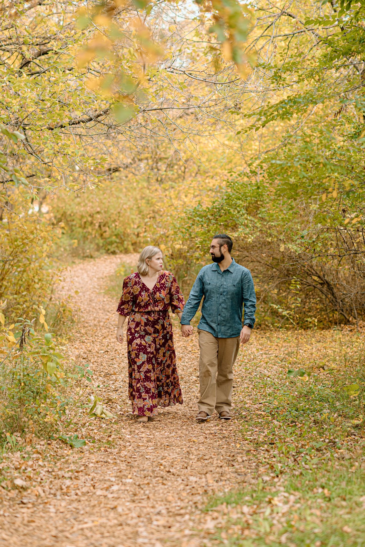 Couple holding hands walking on a leaf-covered path surrounded by golden autumn foliage.