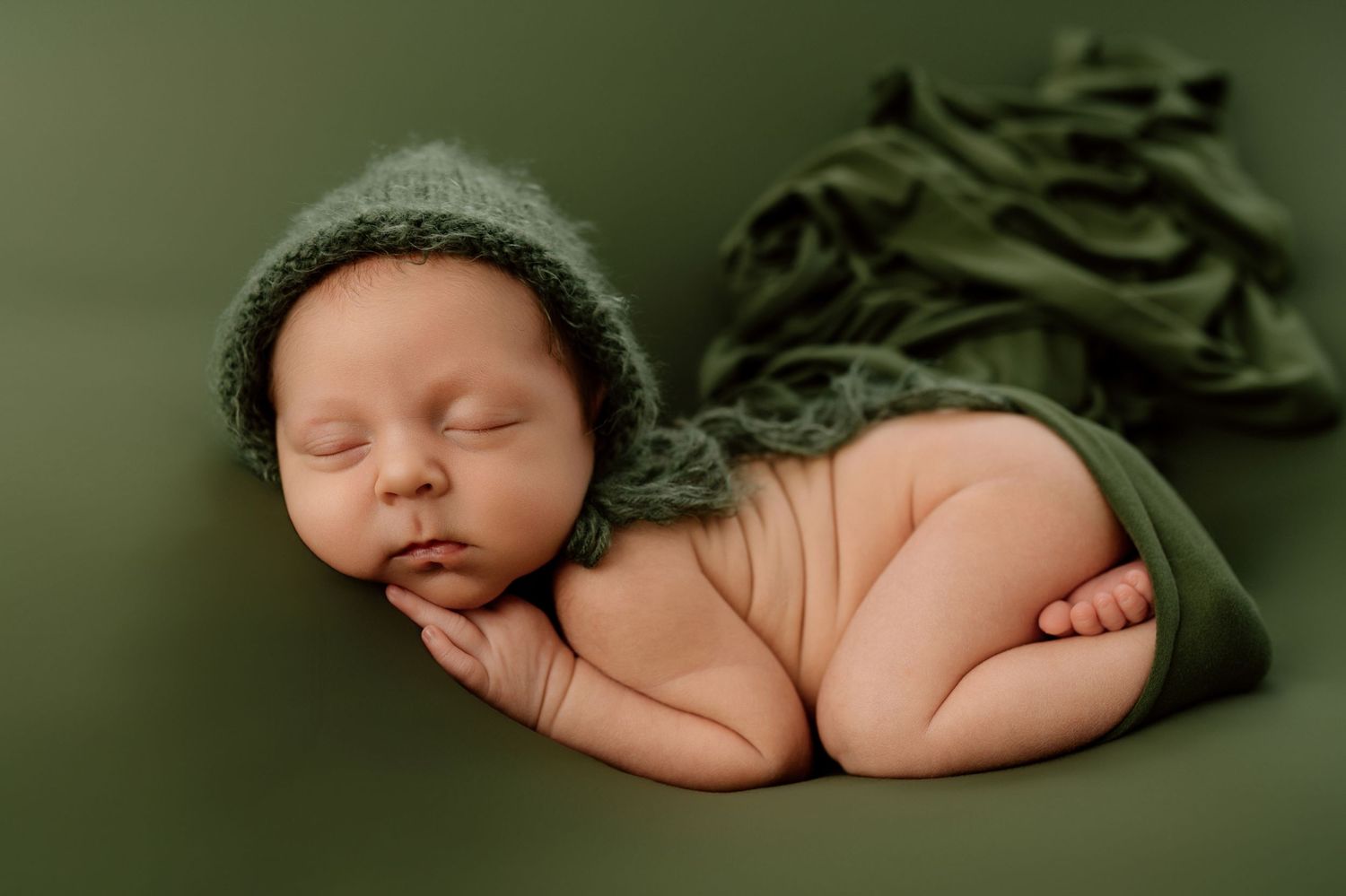 Peaceful newborn baby wearing green knit bonnet sleeping on olive green blanket in studio setting.