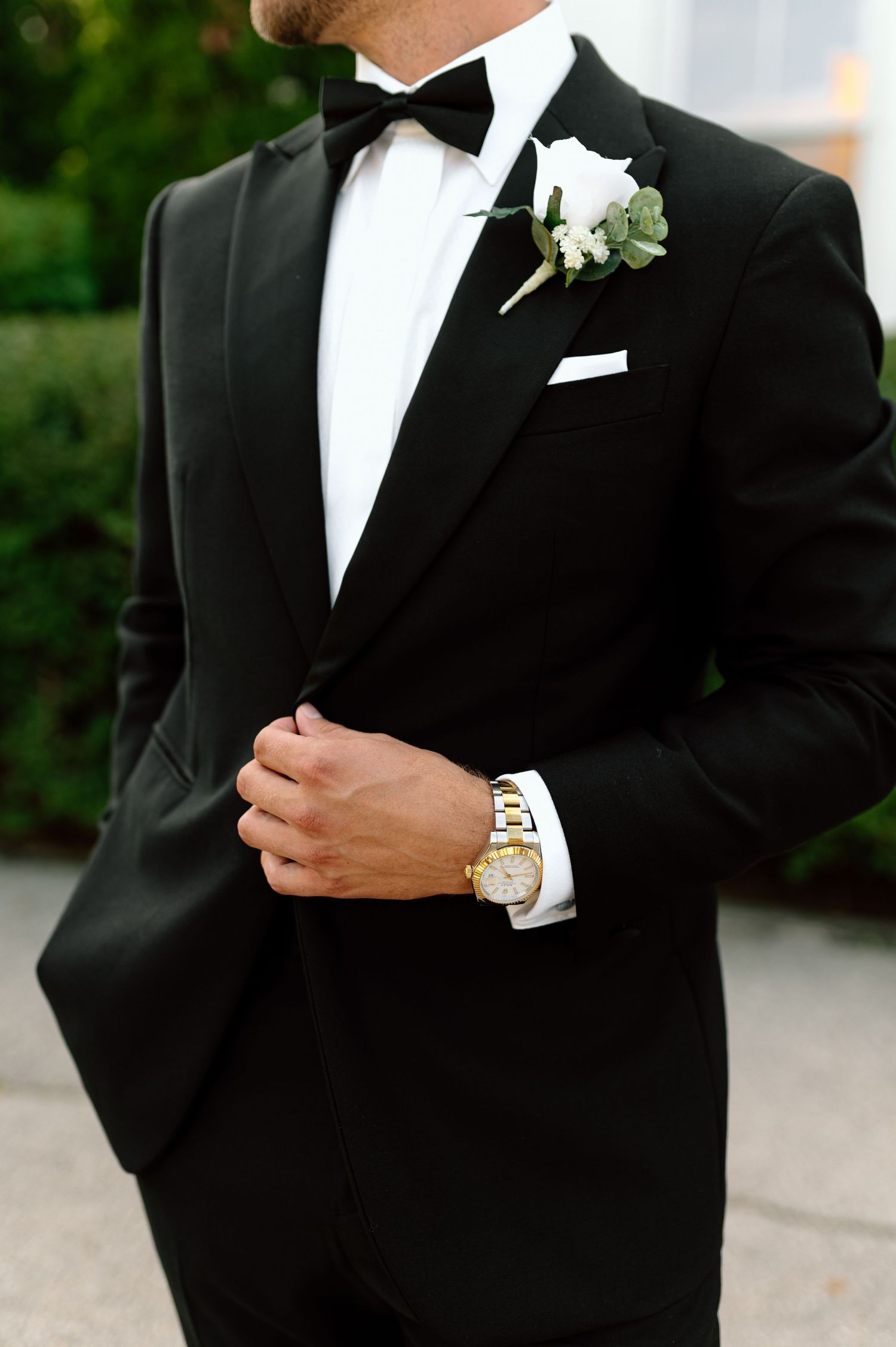 Close-up of groom in black tuxedo with white boutonniere and gold watch at wedding.