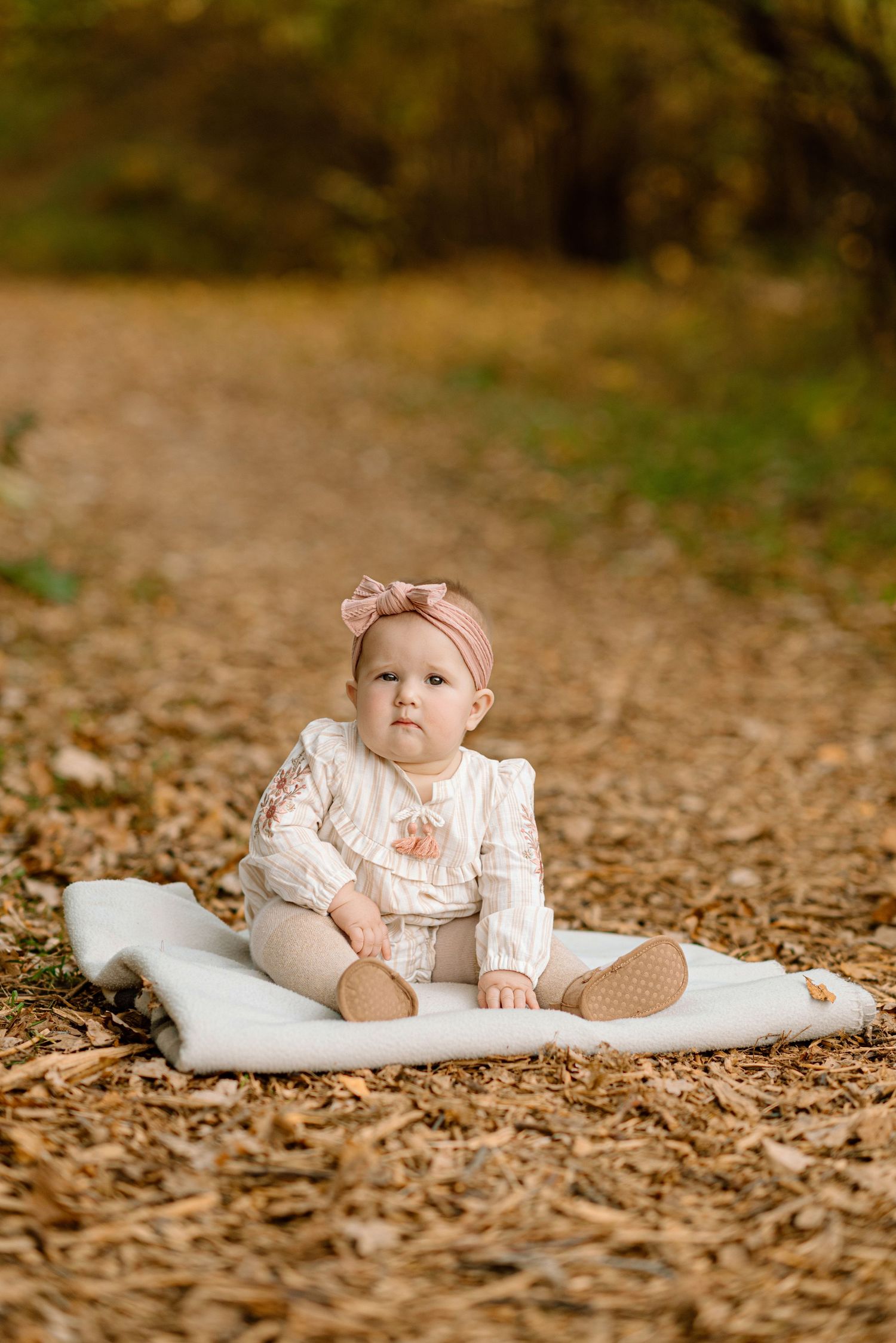 Baby girl in white outfit with pink bow headband sitting on blanket outdoors.