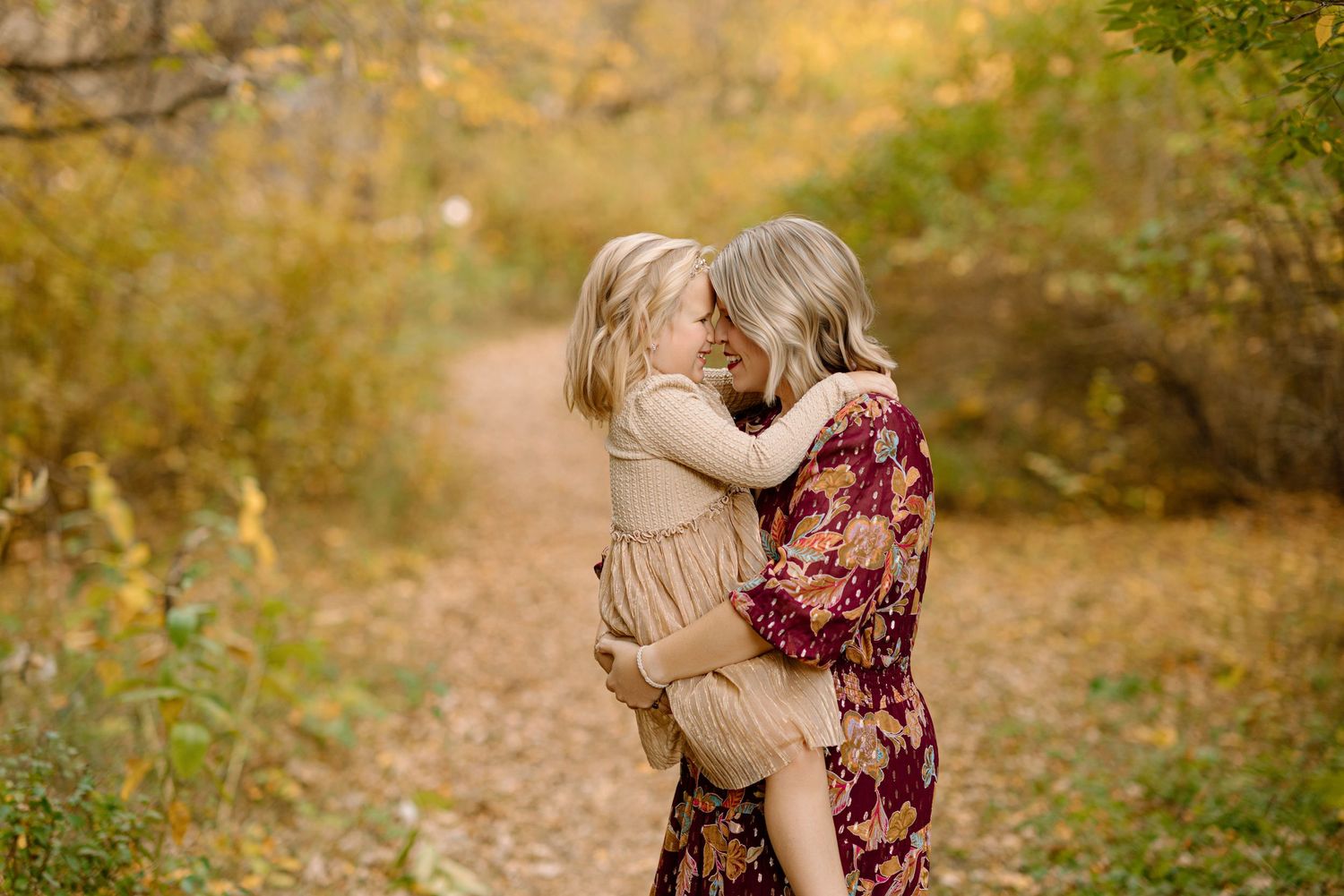 A mother in a floral dress holds her toddler daughter, sharing a tender nose-to-nose moment on an autumn forest path.