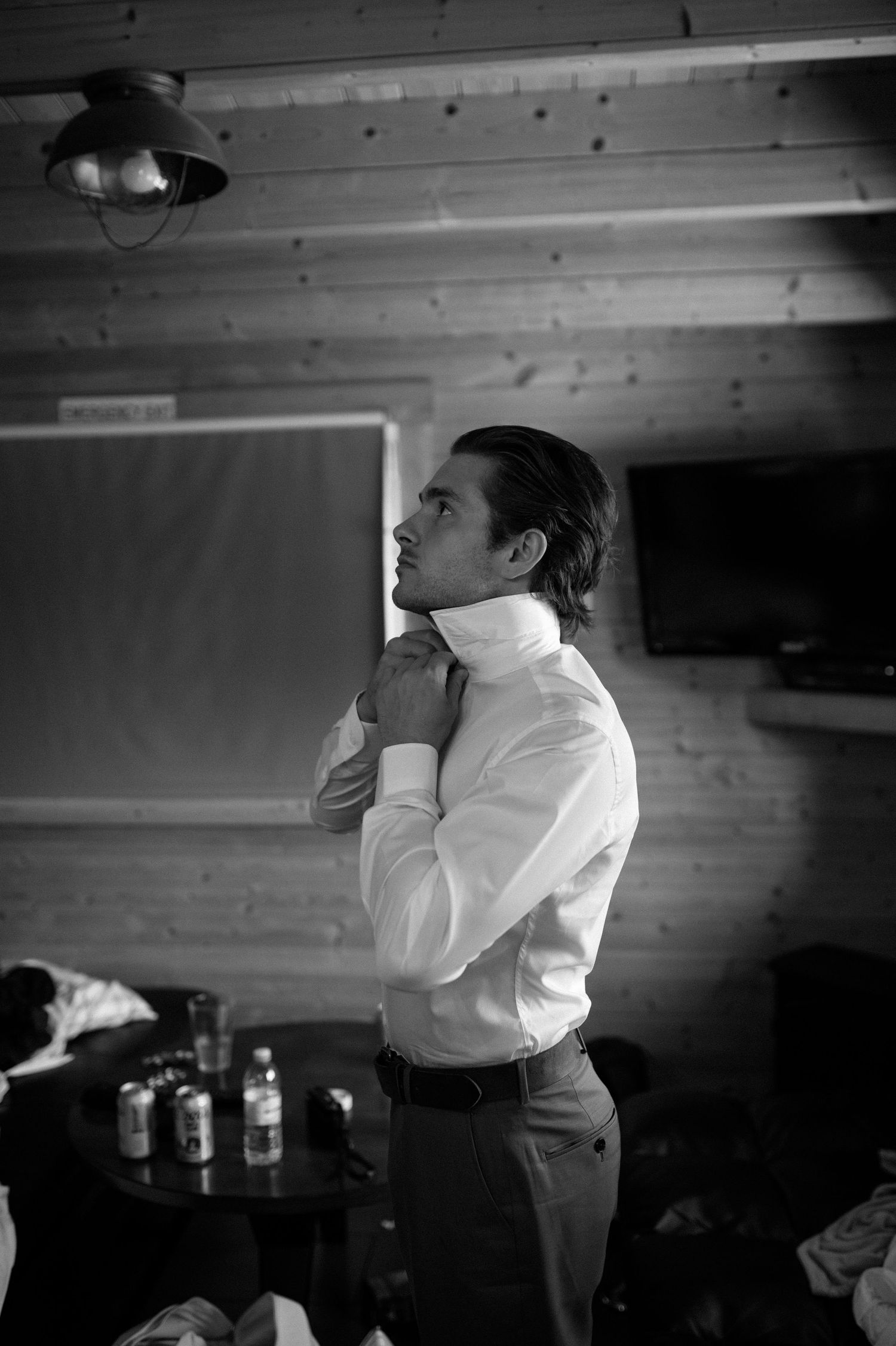 Groom in white dress shirt adjusting collar in rustic wooden room in black and white photo.